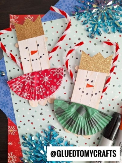 Two handmade king crafts made from craft sticks, gold glitter paper crowns, red and green cupcake liners as skirts, and drawn faces. Perfect companions for a snowman ballerina craft, they rest on festive snowflake-patterned paper with supplies around.