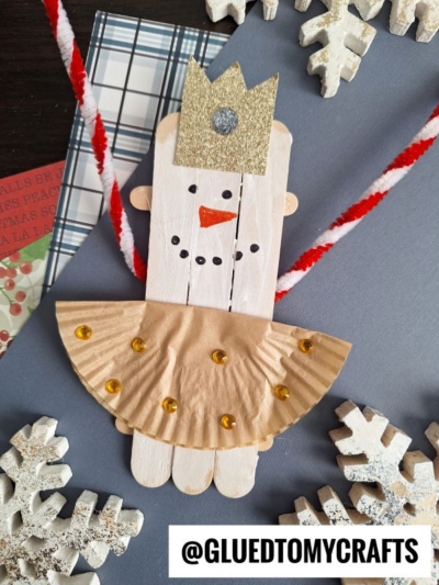 A craft princess made from popsicle sticks and a gold cupcake liner skirt, with a gold glitter crown and sequin decorations, surrounded by white snowflake decorations—perfect to display alongside your favorite popsicle stick snowman ballerina craft.
