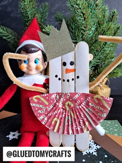 A festive elf doll sits behind a popsicle stick snowman ballerina, featuring gold pipe cleaner arms, a gold crown, candy cane-patterned skirt, and a smiling face. The scene is adorned with greenery and snowflakes.