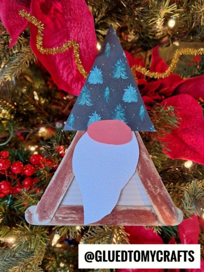 A handmade Popsicle Stick Reindeer Gnome ornament, crafted from painted craft sticks, paper, and glittery material, hangs on a Christmas tree adorned with red poinsettias, berries, and lights. The gnome features a blue beard and a hat with green trees.
