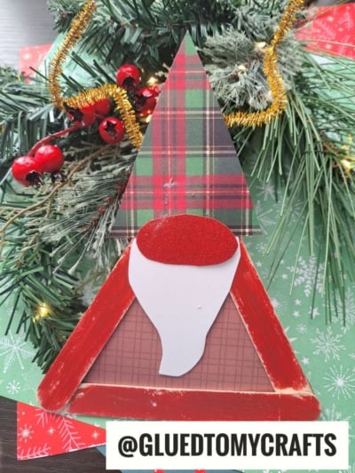 A triangular wooden Christmas gnome with a plaid hat, red body, and white beard sits in front of a festive green background with pine branches and gold tinsel—perfect alongside a Popsicle Stick Reindeer Gnome. Text reads @GLUEDTOMYCRAFTS.