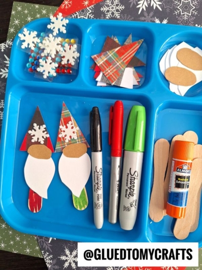 A blue tray with craft supplies: wood sticks, glue, markers, and paper cutouts of gnomes, snowflakes, and other shapes—perfect for creating Christmas gnome scoop stick puppet crafts. The Instagram handle @GLUEDTOMYCRAFTS is at the bottom.