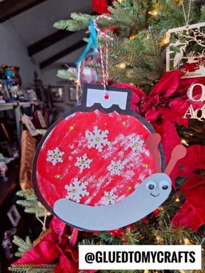 A handmade Christmas ornament snail shaped like a red bauble with white snowflakes and a smiling gray snail, hanging on a decorated Christmas tree. The handle @GLUEDTOMYCRAFTS is displayed at the bottom.