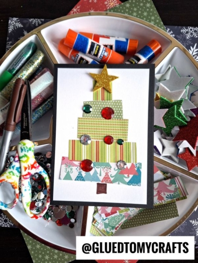 A handmade Christmas card featuring a paper scrap Christmas tree, sequins, and a gold star sits on a tray of craft supplies like glue sticks, scissors, glitter, and paper scraps. The handle @GLUEDTOMYCRAFTS appears at the bottom.