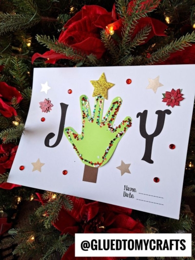 A handprint joy keepsake displaying the word JOY, with a green handprint as the O, decorated with colorful dots, stars, and flowers, hangs on a Christmas tree. There's a name and date space at the bottom right.