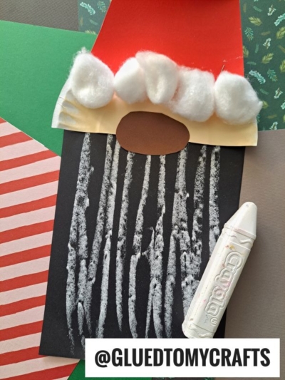 A Chalk Art Santa craft made with colored paper, cotton balls for the hat, white crayon lines for the beard, and @GLUEDTOMYCRAFTS at the bottom. Various patterned papers create a festive background.