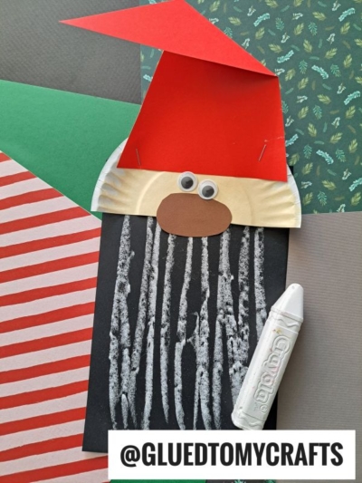 A paper gnome craft with a red hat, googly eyes, and a brown nose sits on a black sheet with white chalk lines, capturing the whimsical charm of Chalk Art Santa. Next to the gnome is a white Crayola crayon, while the background is filled with colored and patterned papers.