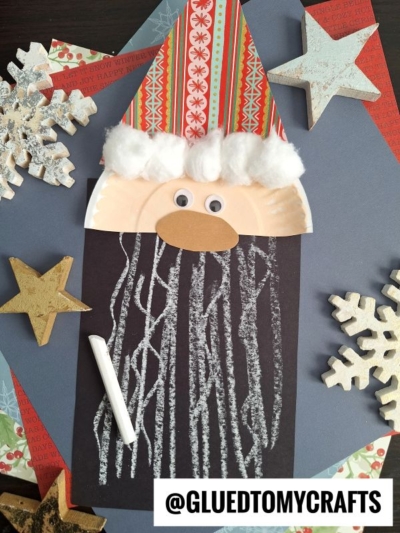 A Chalk Art Santa craft made with a paper plate face, red and patterned paper hat, cotton ball trim, googly eyes, a tan paper nose, and a black beard drawn with white chalk. Snowflake and star decorations surround it. @GLUEDTOMYCRAFTS.