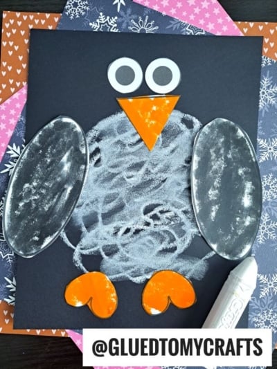 A charming chalk and paper penguin craft made with white chalk on black paper, shiny silver wings, orange feet and beak, and big white eyes. Colorful patterned paper forms the backdrop, with a white crayon in the bottom right corner.