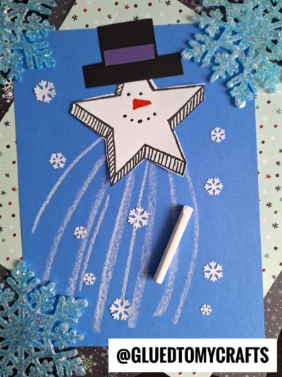 A star-shaped paper snowman with a hat sits on blue paper, surrounded by chalk lines, snowflake stickers, and sparkling blue glitter snowflakes in the corners—almost like a whimsical snowman shooting star racing across a winter sky. A piece of chalk lies on the sheet. @GLUEDTOMYCRAFTS is written at the bottom.