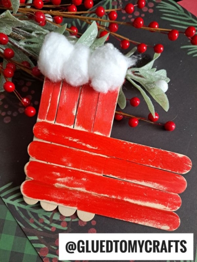 A popsicle stick Christmas stocking, painted red and decorated with white cotton balls and green leaves, is displayed on a festive background with red berries. @GLUEDTOMYCRAFTS is written at the bottom.