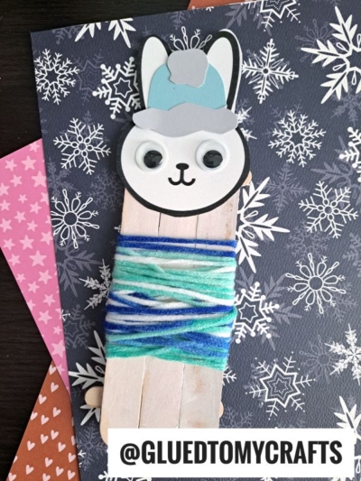 A craft stick snow bunny made from popsicle sticks, blue and white yarn, googly eyes, and a blue winter hat sits on snowflake-patterned paper. Text at the bottom reads @GLUEDTOMYCRAFTS.
