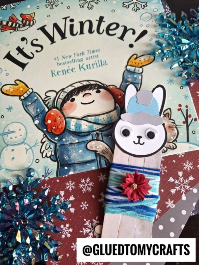 A children's book titled Its Winter! is in the background, with a popsicle stick snow bunny craft wrapped in blue yarn in front. Snowflake decorations and the text @GLUEDTOMYCRAFTS are also visible.