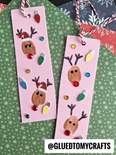Two handmade pink thumbprint reindeer bookmarks decorated with colorful rhinestones, glittery holiday lights, and red, white, and green accents. Each bookmark features red and white string loops at the top.