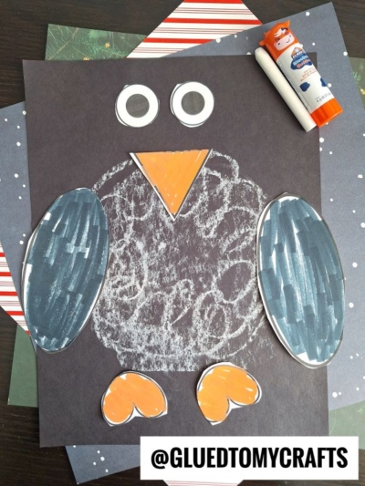 A fun chalk penguin craft made with black paper, white crayon, and colorful cut-out pieces for wings, beak, feet, and eyes. Arranged on a dark background with a glue stick nearby. @GLUEDTOMYCRAFTS is at the bottom.