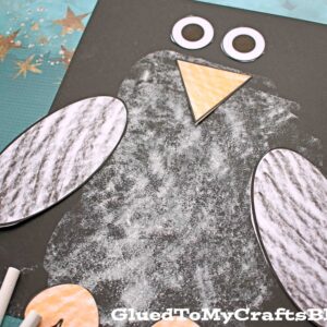 A charming paper & chalk penguin craft made from black paper, white chalk, and cut-out shapes for wings, beak, eyes, and feet is displayed on a blue background with star patterns. Chalk sticks are placed nearby.