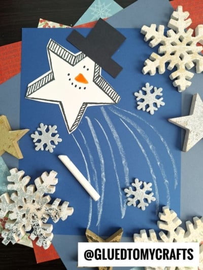 A star-shaped snowman with a hat is glued to blue paper, surrounded by paper snowflakes and wooden stars. Chalk lines suggest a snowman shooting star in motion. A piece of white chalk is nearby. @GLUETOMYCRAFTS is at the bottom.