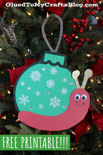 A handmade Christmas ornament snail shaped like a snail, with a green snowflake-patterned shell, gold glittery antennae, and a smiling face, hanging on a festive tree adorned with red berries.