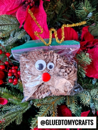 A clear zip-top bag filled with shredded brown crinkle paper, decorated with googly eyes, a red pom-pom nose, and a gold pipe cleaner antler to create a charming crinkle paper reindeer, hangs on a Christmas tree. @GLUEDTOMYCRAFTS is written below.