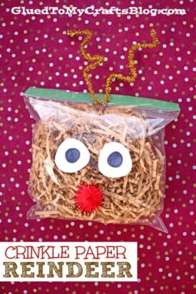 A plastic ziplock bag filled with brown crinkle paper reindeer features googly eyes, a red pom-pom nose, and gold curly pipe cleaner antlers, all set against a red polka dot background.