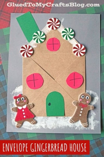 A paper envelope gingerbread house made from a brown envelope, with red and white peppermint circles, red windows, green door and roof, plus two paper gingerbread people, on a gray background with Envelope Gingerbread House label.