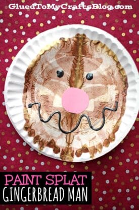 A paint splat gingerbread man face decorates a paper plate, featuring a pink paper nose, googly eyes, and a wavy black mouth, all set against a cheerful red polka dot background.