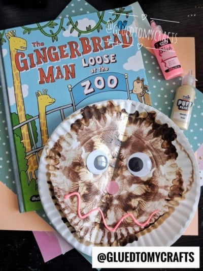 A paint splat gingerbread man made from a paper plate with big googly eyes sits on a colorful background next to the book The Gingerbread Man Loose at the Zoo and bottles of puffy paint.