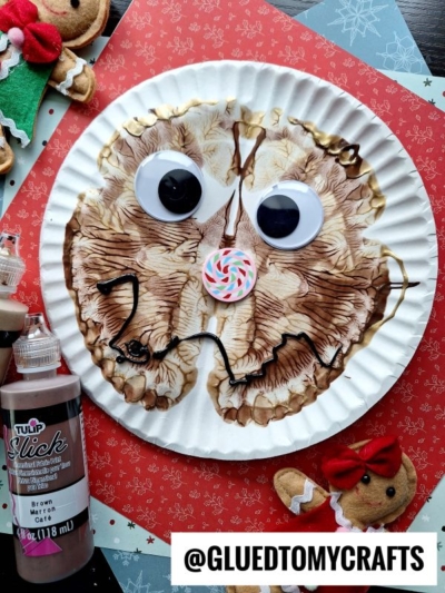 A paper plate reindeer face with brown paint, large googly eyes, and a colorful candy nose sits among red patterned paper, paint bottles, a gingerbread man, and a festive paint splat. @GLUEDTOMYCRAFTS appears at the bottom.