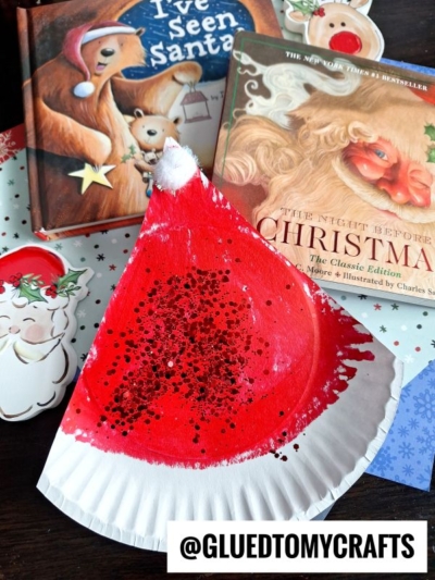 A red and white paper plate Santa hat craft with glitter is displayed in front of Christmas books and festive decorations, alongside a playful paper plate elf hat. The Instagram handle @GLUETOMYCRAFTS is at the bottom of the image.