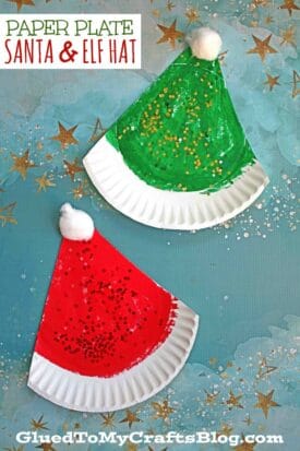 Two paper plates are transformed into a paper plate Santa hat, painted red with a white pom-pom and glitter, and a paper plate Elf hat, painted green with gold glitter for festive holiday fun.