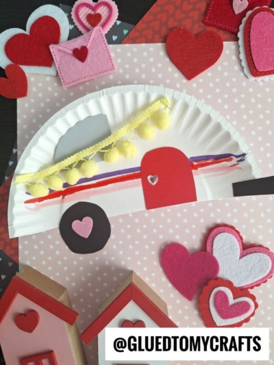 A Valentine-themed collage featuring a paper plate camper craft for kids, felt hearts, a yellow pom-pom garland, houses, and heart decorations on a pink heart-patterned background. The handle @GLUEDTOMYCRAFTS appears at the bottom.