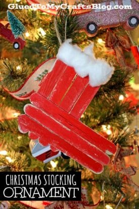 A handmade popsicle stick Christmas stocking ornament with red paint and cotton hangs on a decorated tree adorned with lights, ribbon, and other festive ornaments.