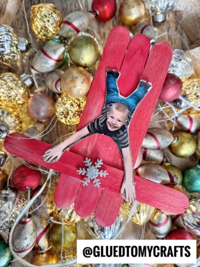 A red popsicle stick sleigh ornament showcases a cut-out photo of a smiling child, surrounded by festive gold and silver ornaments. The sled features a snowflake embellishment, with the handle @GLUEDTOMYCRAFTS at the bottom.