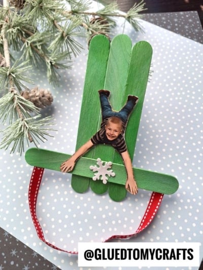 A popsicle stick sleigh ornament crafted from green sticks is shaped like a sled, decorated with a child’s photo cutout, a white snowflake, and a red ribbon loop. Pine branches accent the background. Text reads @GLUEDTOMYCRAFTS.