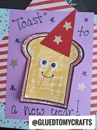 Handmade greeting card on a purple background featuring a slice of toast with googly eyes and a red party hat, with the text Toast to a new year!.