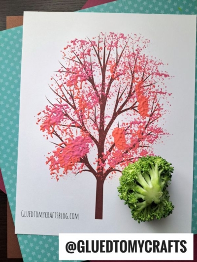 A printed tree on white paper is decorated with pink and orange paint, creating a broccoli stamped cherry blossom effect. The broccoli sits near the bottom right of the page, and colored papers are layered underneath.
