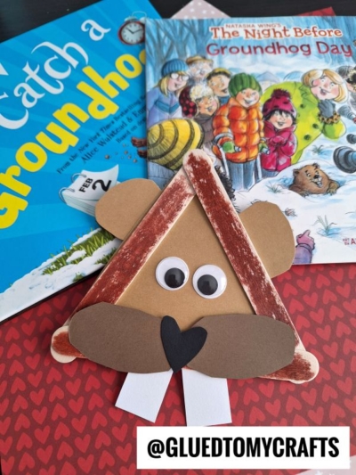 A popsicle stick groundhog craft made from brown paper sits on a red heart-patterned surface, with two children's books about Groundhog Day in the background. The Instagram handle @GLUEDTOMYCRAFTS is visible at the bottom.