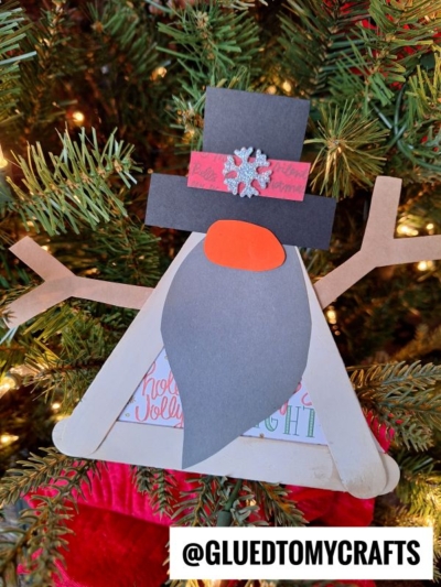 A handmade popsicle stick snowman gnome ornament made from craft sticks, felt, and paper hangs on a Christmas tree. The gnome has a gray beard, orange nose, black hat with a snowflake, and arms. Text at the bottom reads @GLUEDTOMYCRAFTS.
