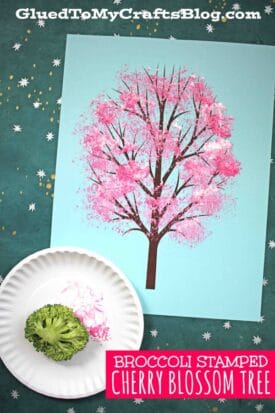 A blue sheet displays a painted cherry blossom tree with pink blossoms. A broccoli floret and a plate of pink paint reveal how this broccoli stamped cherry blossom design was created. BROCCOLI STAMPED CHERRY BLOSSOM TREE is written at the bottom.