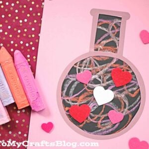 A pink paper cutout love potion bottle filled with colorful paper chalk scribbles, decorated with heart shapes. Three pastel chalk pieces rest nearby on a red, gold-spotted background.