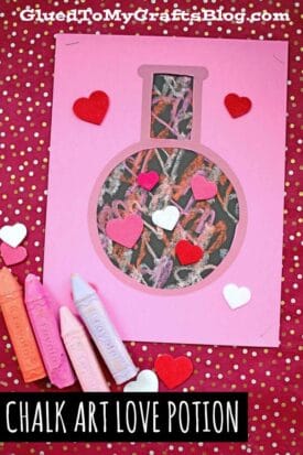 A pink paper cutout love potion bottle filled with colorful paper chalk scribbles, decorated with heart shapes. Three pastel chalk pieces rest nearby on a red, gold-spotted background. Text reads Chalk Art Love Potion.
