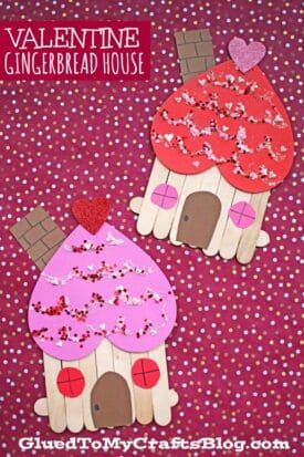 Two valentines gingerbread house crafts made with popsicle sticks, heart-shaped roofs, glitter, and heart decorations, displayed on a red and white polka dot background.