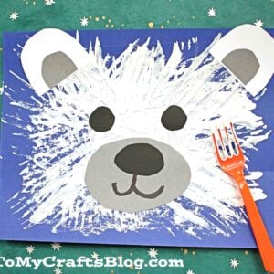 An adorable fork painted polar bear craft done on blue paper rests on a green surface with tiny stars. The bear features white painted fur, gray paper ears and snout, and black paper eyes and nose. An orange fork lies beside the finished bear craft.
