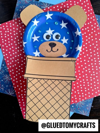 A blue paper plate crafted into an ice cream cone bear’s face with white stars, googly eyes, and a black nose sits atop a paper cone on red and blue star-patterned paper. The image is labeled @GLUEDTOMYCRAFTS.