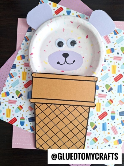 A cute ice cream cone bear crafted from paper, featuring a bear face with googly eyes, paper ears, and a drawn nose and mouth on a plate, displayed atop colorful patterned paper. @GLUEDTOMYCRAFTS is written at the bottom.