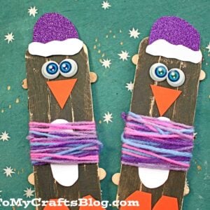 Two popsicle stick penguins with googly eyes, orange beaks, purple hats, and colorful yarn scarves look like adorable penguins in Christmas sweaters, set on a green background with white stars.