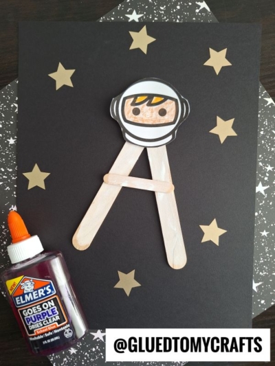 A black paper with gold stars displays a popsicle stick letter A decorated as an astronaut for an "A is for Astronaut" craft. Elmers purple glue sits on the left, and @GLUEDTOMYCRAFTS is written at the bottom right.