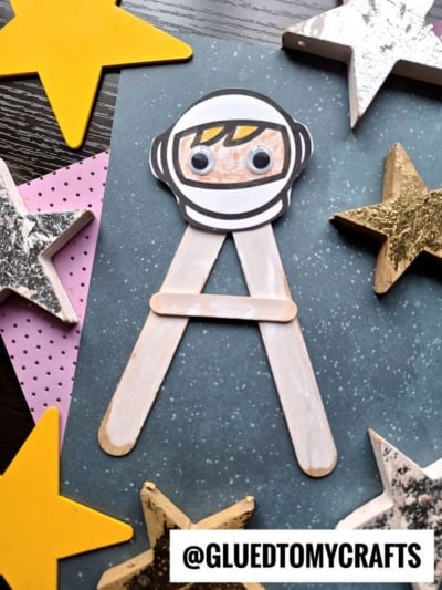 A is for astronaut in this space-themed craft, featuring a popsicle stick astronaut with a paper helmet and googly eyes, surrounded by yellow and gold stars on a blue, starry background. Username @GLUEDTOMYCRAFTS is at the bottom.