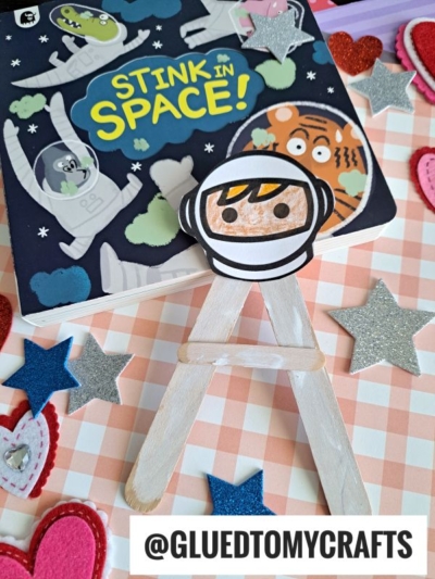 A children’s book titled Stink in Space! sits on a checkered surface, surrounded by felt hearts and glittery stars. In front is a handmade “A is for Astronaut” craft on popsicle sticks. @GLUEDTOMYCRAFTS is displayed at the bottom.