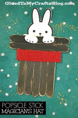A popsicle stick magician's hat craft features black sticks, a red glittery band, and a cute white bunny peeking out. The teal background is sprinkled with white star shapes for extra charm.